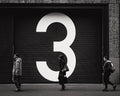Photo noir et blanc d’une porte de garage avec le chiffre 3 peint en grand, devant laquelle trois personnes marchent dans une rue de La Haye