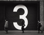 Photo noir et blanc d’une porte de garage avec le chiffre 3 peint en grand, devant laquelle trois personnes marchent dans une rue de La Haye