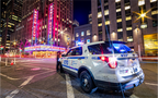 Photo de nuit à New York avec un véhicule du NYPD aux feux allumés au premier plan et le Radio City Music Hall en arrière-plan, ambiance lumineuse urbaine.
