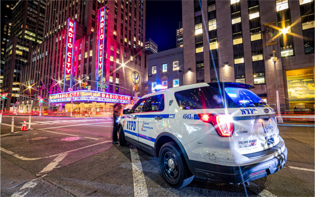 Photo de nuit à New York avec un véhicule du NYPD aux feux allumés au premier plan et le Radio City Music Hall en arrière-plan, ambiance lumineuse urbaine.