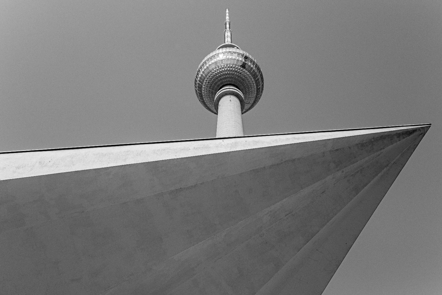 Photo noir et blanc de la tour Fernsehturm à Berlin vue en contre-plongée, avec la pointe de la tour apparaissant derrière un bâtiment à l’architecture angulaire