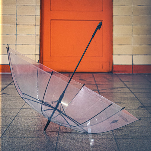 Photo d’un parapluie transparent ouvert posé au sol dans l’entrée d’une station de métro à Berlin, devant une porte orange, ambiance vintage et urbaine