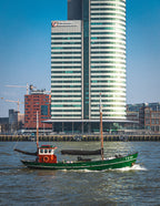 Photo d’un voilier vert naviguant devant un building moderne à Rotterdam, illustrant un contraste entre nature et architecture urbaine